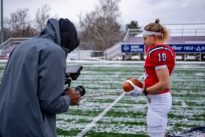 Haley Van Voorhis in a red ĻӰ football uniform holding a football on a football field while a videographer records footage.